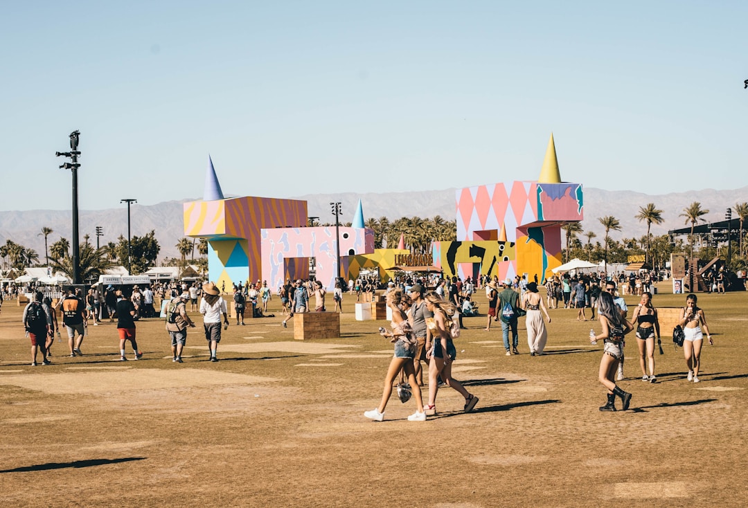 A lively outdoor festival scene with people walking and gathering in an open field. Colorful geometric structures with artistic designs are prominently displayed in the background amidst palm trees. The atmosphere is bright and sunny, indicative of a warm day.
