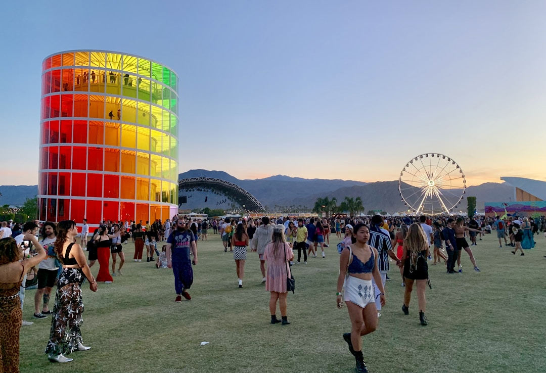A vibrant outdoor festival scene with people walking and gathering on a vast grassy area during sunset. A tall, cylindrical structure with colorful panels stands prominently on the left. A large Ferris wheel is visible in the background against a backdrop of mountains. Attendees are casually dressed and engaged in various activities.