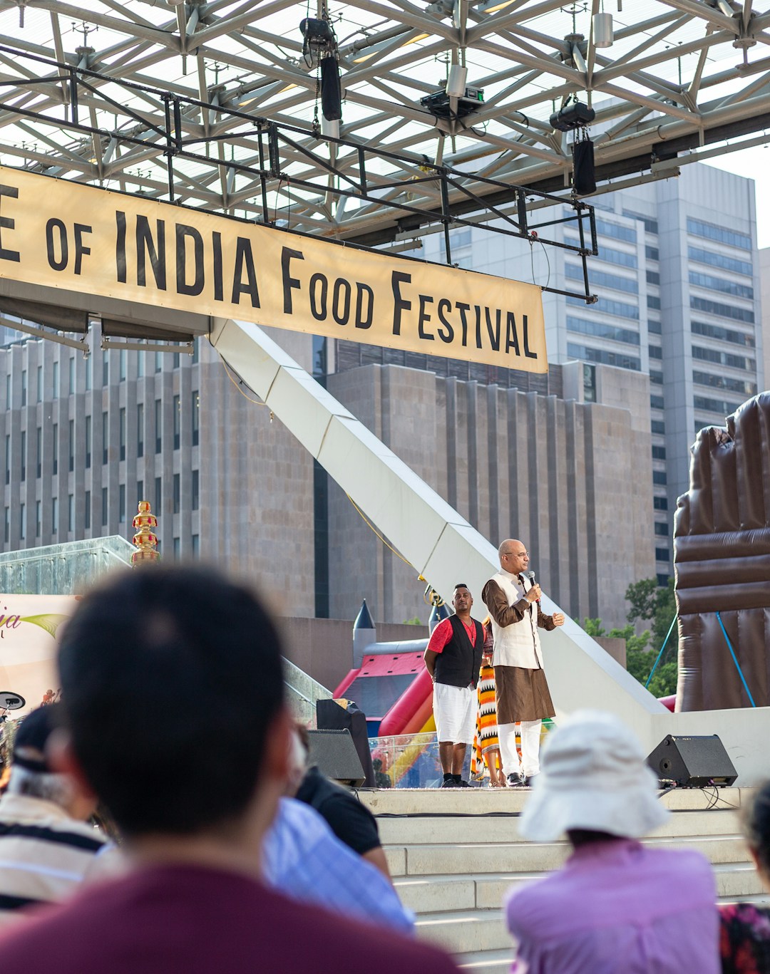 A food festival with a sign reading 'Taste of India Food Festival' takes place outdoors. There is a stage with a person speaking into a microphone, surrounded by attendees. Musical performers with traditional instruments are present. Surrounding the event are modern buildings and an inflatable structure.