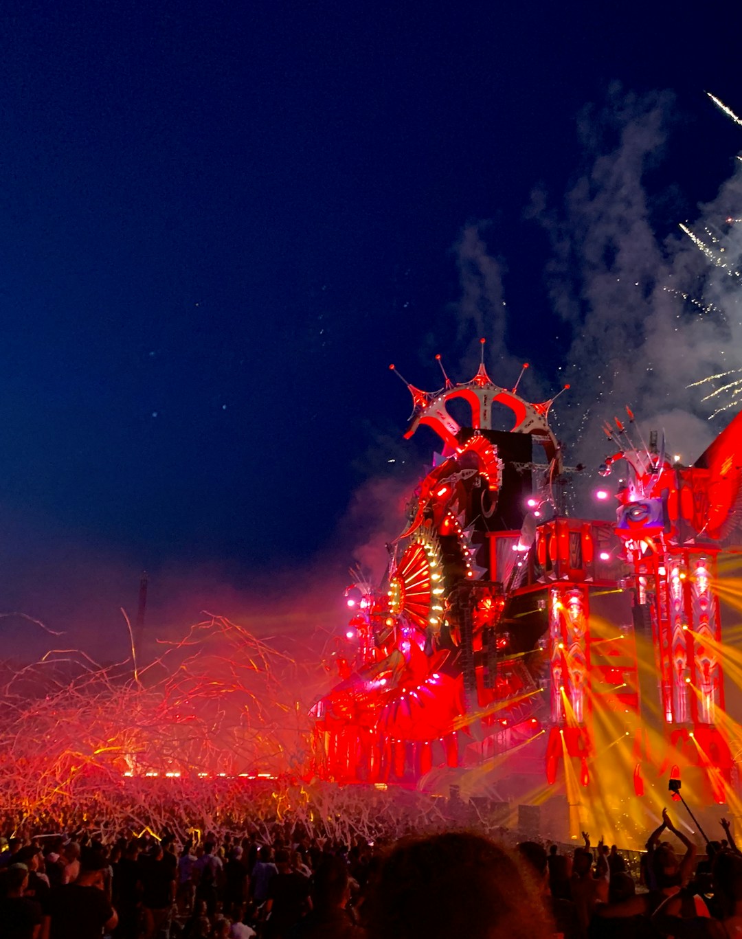 An energetic outdoor festival at night with a large crowd gathered in front of an elaborate stage adorned with vibrant red and orange lights. Fireworks explode in the night sky, adding to the lively atmosphere.