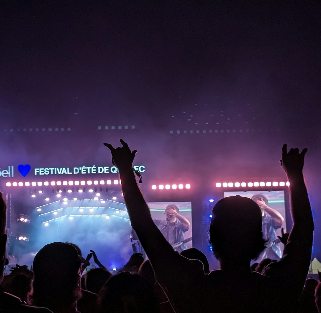 A large outdoor music festival at night features a stage with bright lights and a video screen displaying a musician. People in the crowd are silhouetted against the colorful lights, some with their arms raised in excitement.
