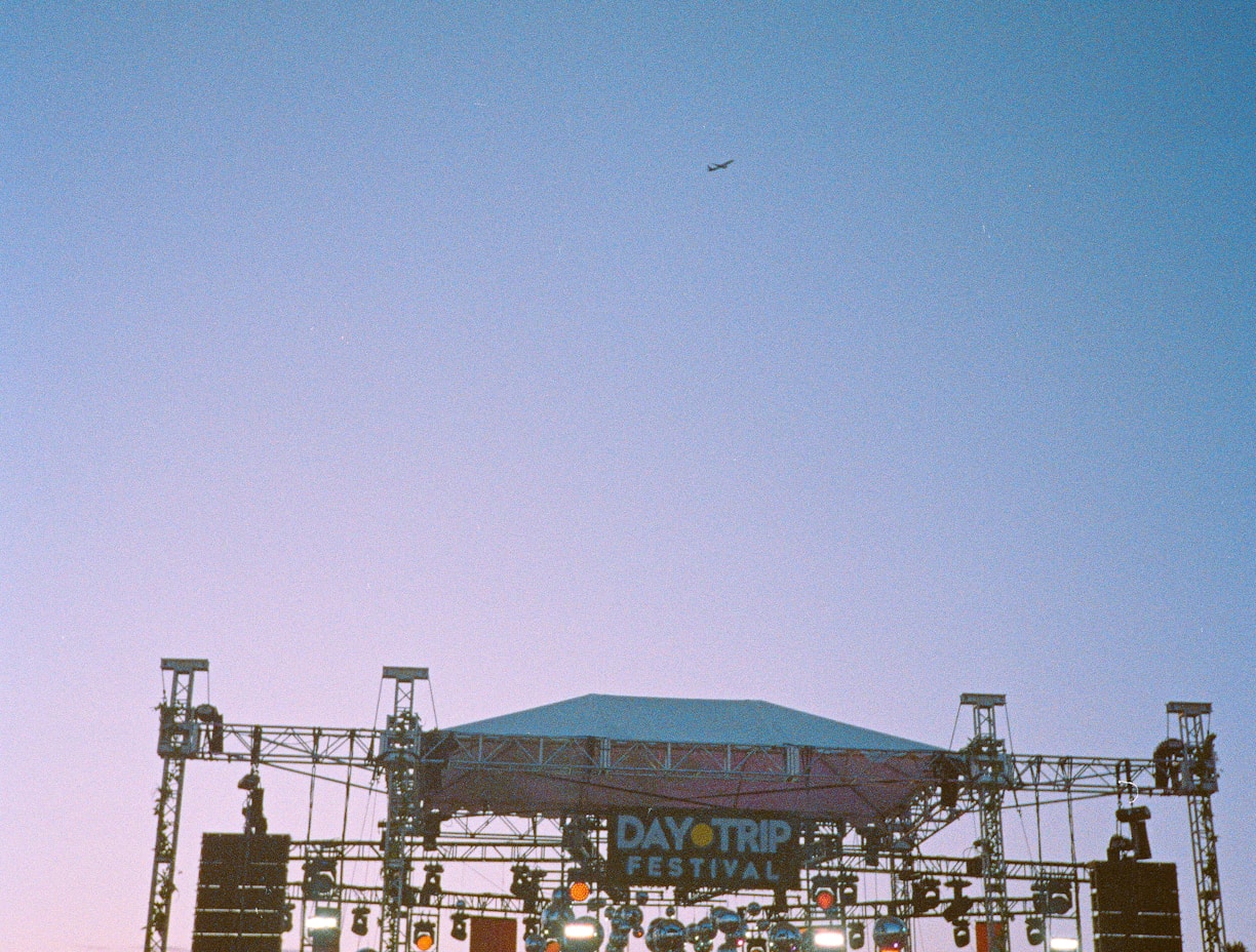 A large outdoor music festival during sunset with a stage set up featuring lights and decorations. A crowd of people in hats and vibrant clothing is gathered in front of the stage, enjoying the performance. The sky above is clear with a hint of an airplane in the distance.